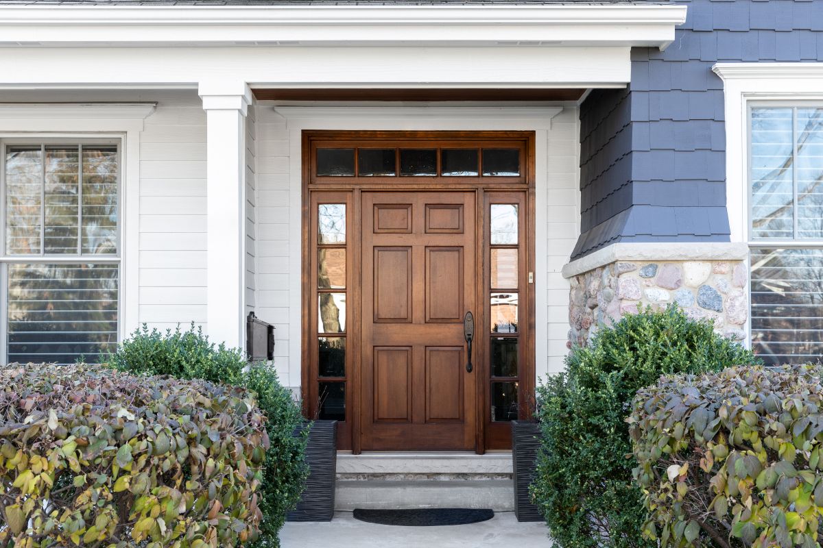 A front door and covered porch detail on a home with grey siding, white trim, red brick, a wooden front door.